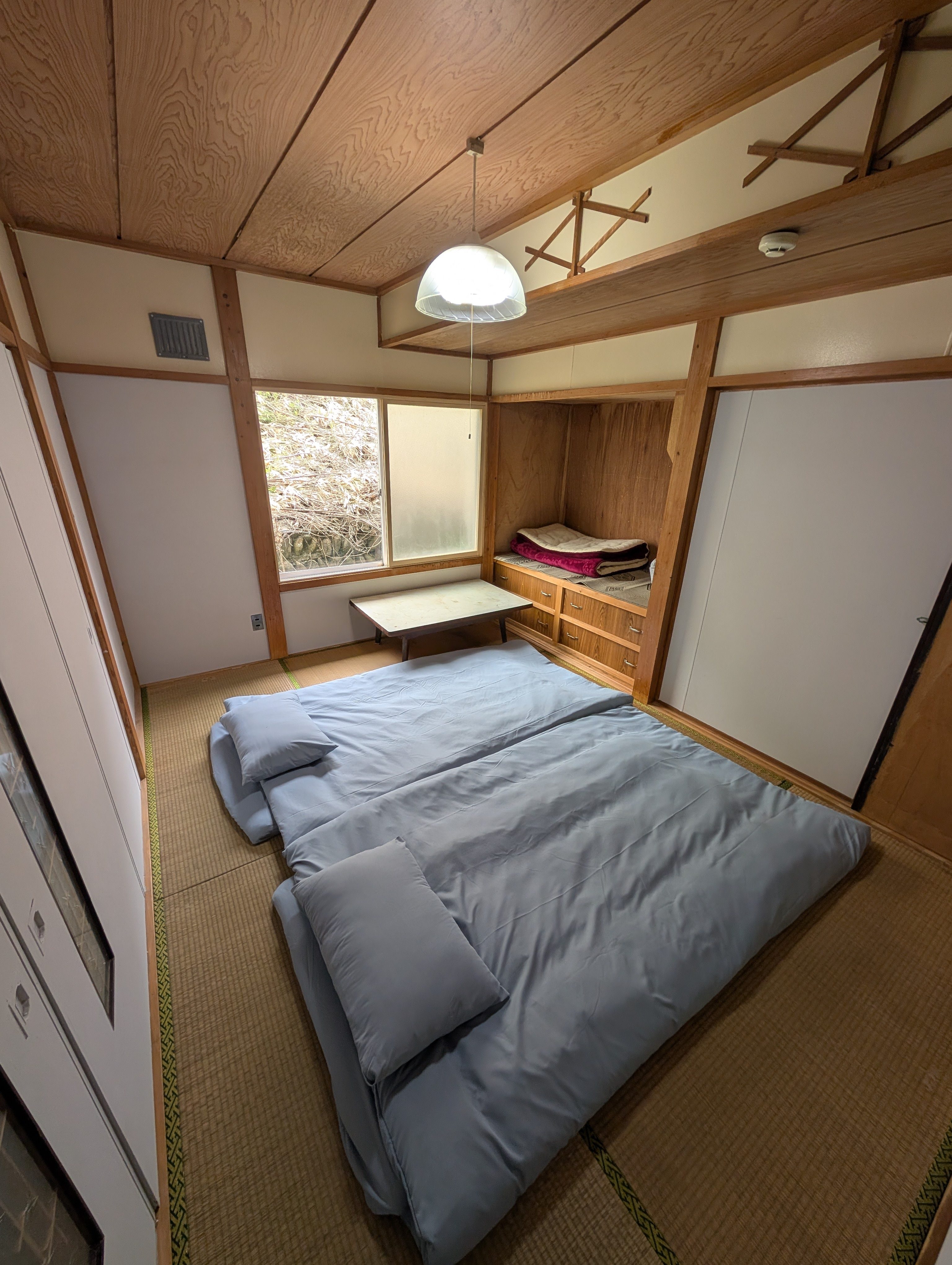 木の天井と低いテーブル、すりガラスの窓のそばに布団を敷いた1階の畳の部屋。